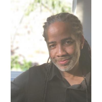 A picture of Jonathan W. Gray sitting in front of a window. He's an African American male wearing a dark shirt with his dreadlocks tied back. 